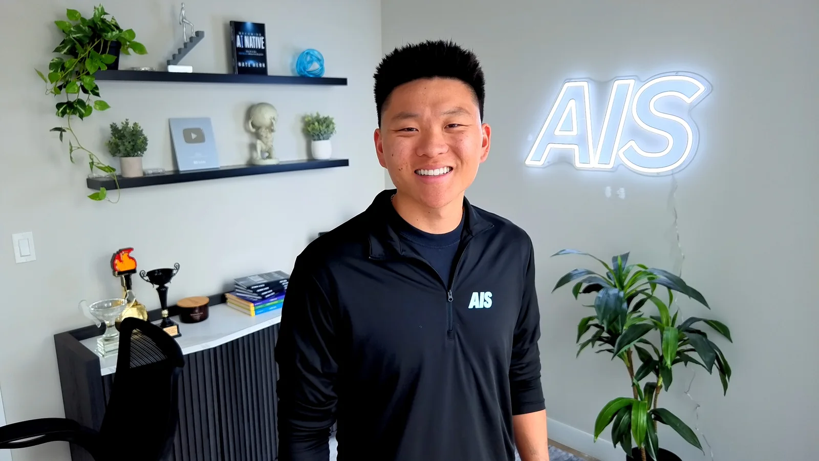 Nate Herk in his YouTube studio, an AIS neon sign behind him.